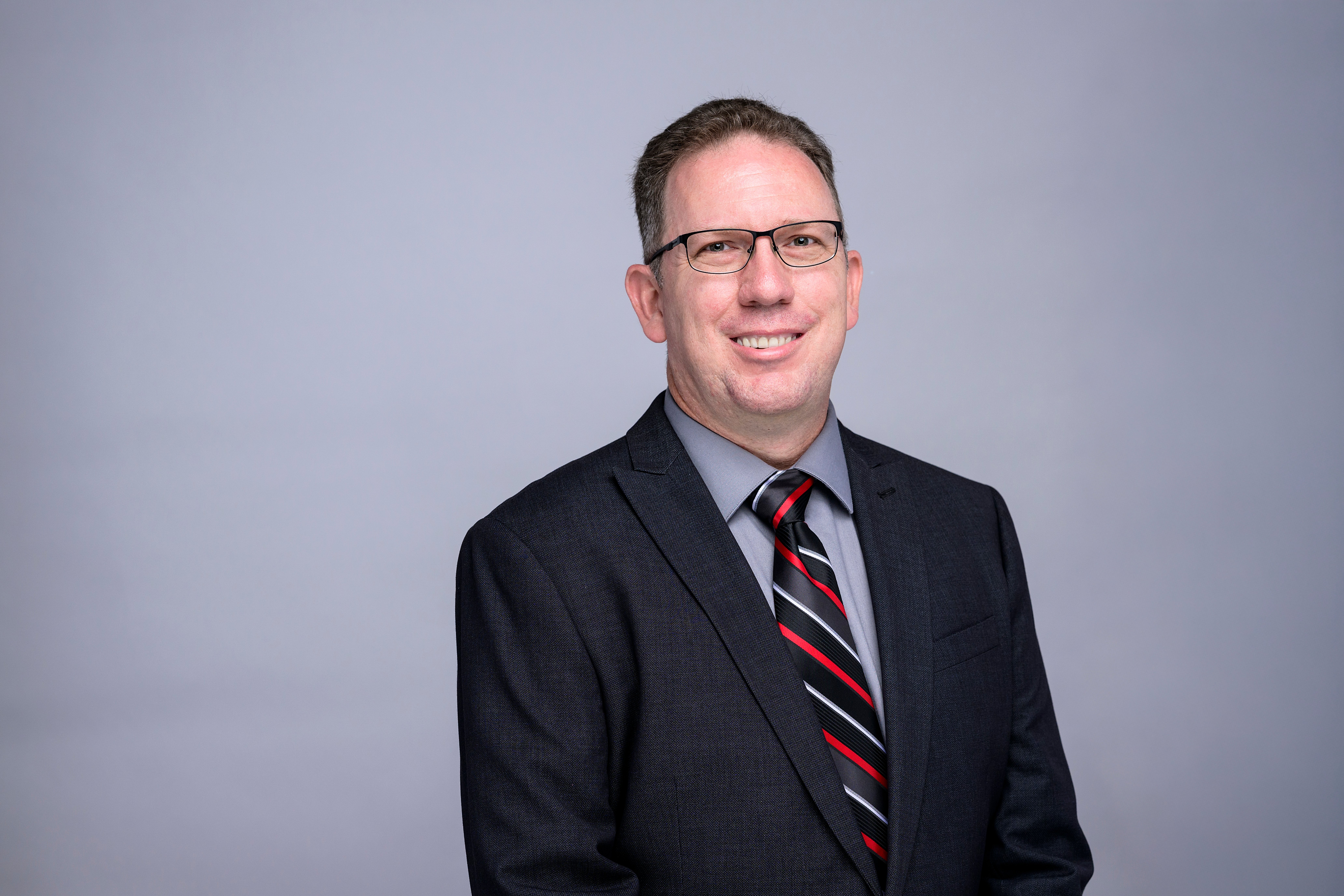 Man with glasses on smiling and a black jacket and striped tie with a grey background, smiling